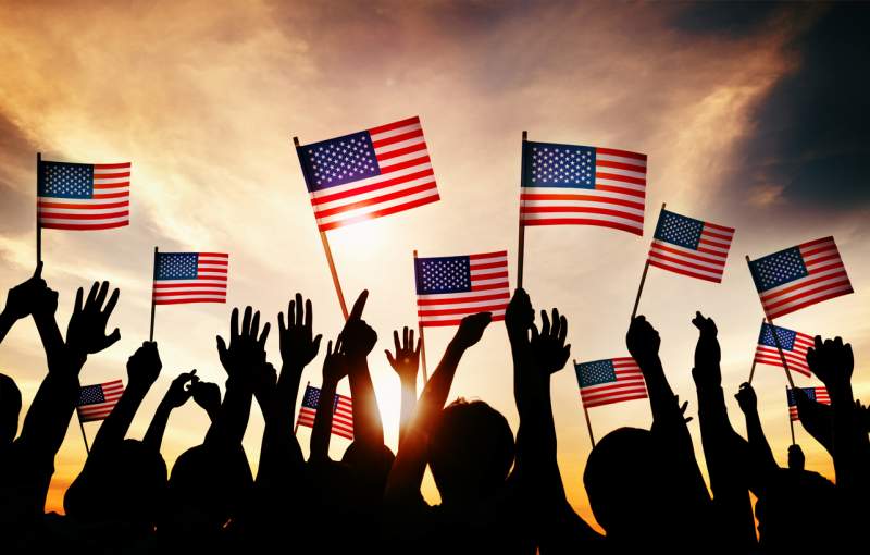 Group of adults and children holding United States flags, facing away and silhouetted by the sun.